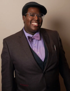 Lianna is smiling at the camera. Lianna is wearing a purple shirt and purple bowtie with a brown jacket, glasses, and a brown flat cap.