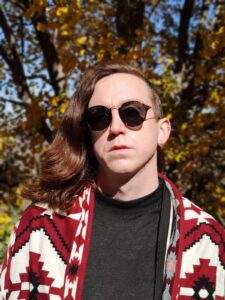 Juani is wearing a red, black, and white pattered jacket over a grey top. She is wearing sunglasses and looking into the camera. In the background is a tree with yellowing leaves.